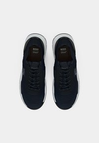Paire de chaussures de sport BOSS noires et blanches avec lacets et semelle texturée, vue de dessus sur un fond clair uni.