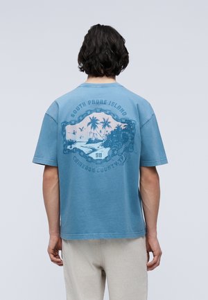 Person mit dunklem Haar trägt ein blaues T-Shirt mit einer Grafik einer tropischen Insel und dem Text "South Padre Island" und "Cameron County, TX" auf der Rückseite.