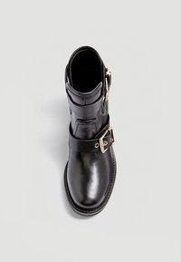 Botte cheville en cuir noir avec un bout rond, présentée avec deux accents de boucle en or et une sangle texturée autour de la cheville. Fini lisse.