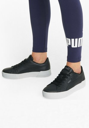 Baskets en cuir noir avec une surface texturée, semelle en caoutchouc blanche et logo Puma bien visible sur le côté, associées à des leggings navy.
