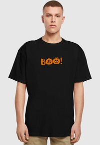 Must t-särk, mis on valmistatud puuvillast, esitleb oranži "Boo!" teksti koos kahe kõrvitsa näoga. Lahtine lõige, lühikesed varrukad, lihtne disain.