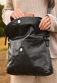 Una borsa nera in pelle con una texture liscia, dotata di un ribaltina curva e due chiusure a scatto in argento. Un portafoglio color cammello è parzialmente visibile all'interno.