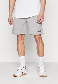 Pantalones cortos de algodón grises con bolsillos laterales, con un dobladillo enrollado y un logo de "ellesse" en la parte inferior izquierda. Llevados con calcetines blancos y zapatillas.