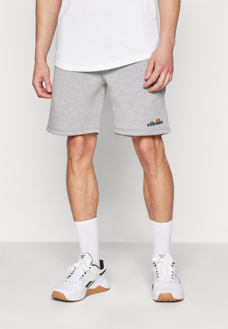 Pantalones cortos de algodón grises con bolsillos laterales, con un dobladillo enrollado y un logo de "ellesse" en la parte inferior izquierda. Llevados con calcetines blancos y zapatillas.