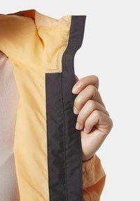 Mano sosteniendo el borde de una chaqueta amarilla con ribete negro y forro de malla, mostrando costuras a lo largo de los bordes y una textura suave.