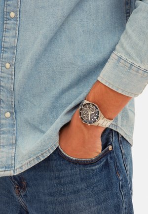 Zilveren roestvrijstalen horloge met een zwarte wijzerplaat, witte markeringen en chronograaf functies, gedragen om een pols met een denim shirt en jeans.