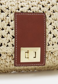 Woven straw handbag z prostokątną brązową skórzaną łatą i złotym metalowym zapięciem o kwadratowym wzorze, ozdobiona białym przeszyciem.
