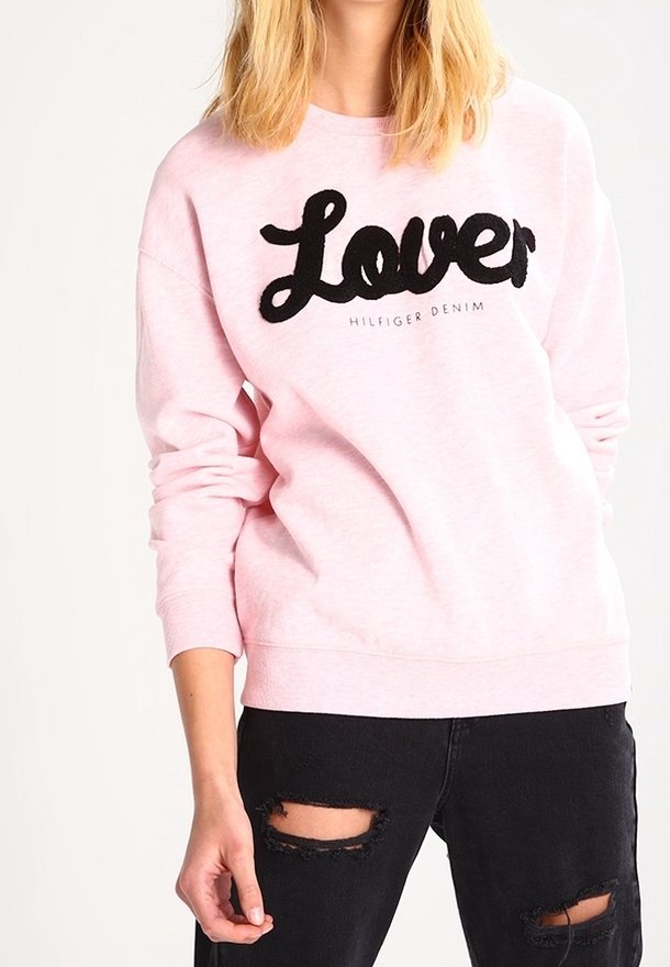 Lichtroze sweatshirt met oversized zwarte "Lover" tekst en "Hilfiger Denim" detail, met geribde manchetten en zoom, gedragen met versleten zwarte jeans.