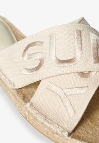 Sandalia de esparto beige con diseño de tiras cruzadas y sutil bordado de marca, suela de yute natural y suave tela texturizada.