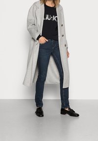 Ljust grå ullkappa med slagkrage, ensidig design, svart tröja med vit logotyp, mörkblå jeans, svarta skor.