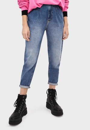 Person trägt hochgekrempelte blaue Jeans, schwarze Schnürkampfstiefel und einen pinkfarbenen Pullover mit schwarzen Bündchen, stehend vor einem schlichten Hintergrund.