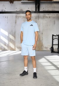 Helle blaue Sport-T-Shirt mit schwarzem Logo auf der Brust, passende hellblaue Shorts, schwarze Sneaker und weiße Socken. Betonhintergrund.
