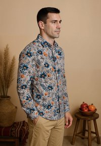 Man in beige broek en een bloeiende lange mouw shirt, staand met zijn handen in zijn zakken in een kamer met neutrale tinten en decor.