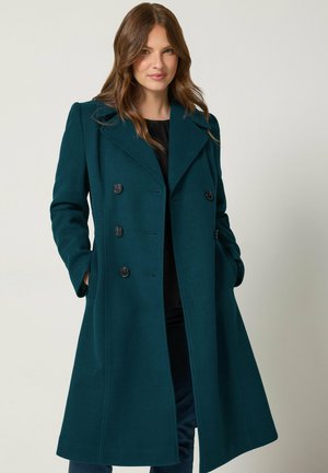 Femme aux longs cheveux bruns portant un manteau croisé bleu sarcelle foncé, les mains dans les poches, sur un fond clair uni.