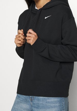 Persoon draagt een zwarte Nike-hoodie met wit swoosh-logo, houdt zwarte trekkoorden vast, gecombineerd met lichtblauwe spijkerbroek.