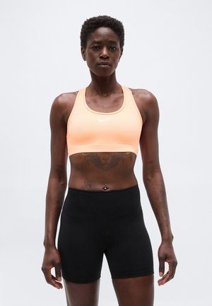 Atletická osoba v broskyňovej športovej podprsenke a čiernych vysokých šortkách stojaca na svetlošedom pozadí.