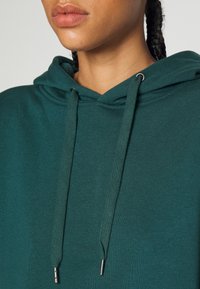 Huvtröja i mörkgrön bomull, med dragsko och silverfärgade metalldelar, samt en mjuk textur. N ärbild på halslinjen.