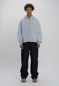 Felpa con zip azzurra chiara, dotata di tasca frontale, abbinata a jeans scuri larghi e calzature color sabbia. Il tessuto appare morbido con una vestibilità comoda.