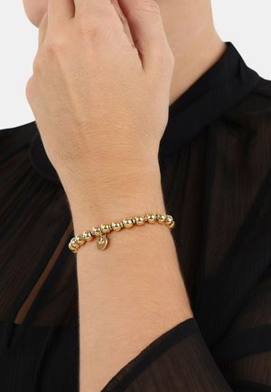 Pulsera de cuentas en tono dorado con un pequeño charm en forma de corazón con "100" grabado. Llevada en una muñeca, sobre un fondo de tela negra transparente.