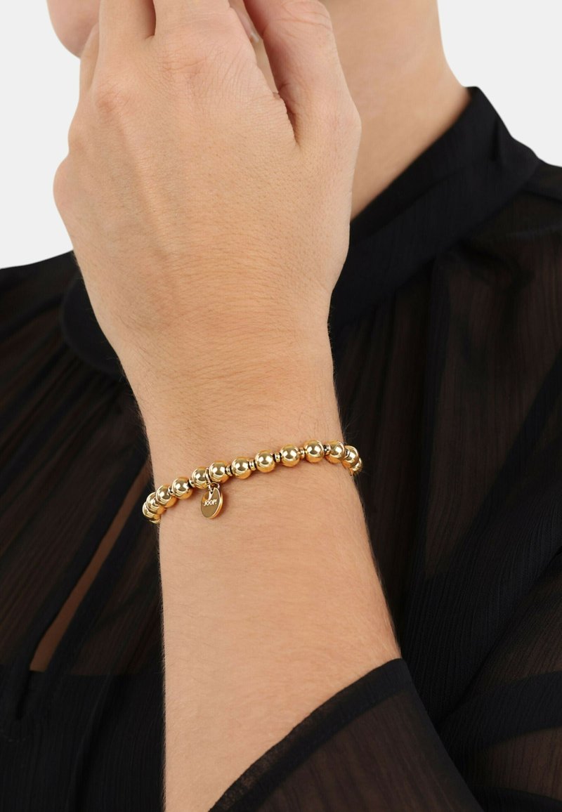 Bracciale in tono oro con perline, dotato di un piccolo ciondolo a forma di cuore con inciso "100". Indossato su un polso, su uno sfondo di tessuto trasparente nero.