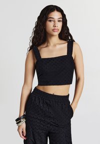 Donna con lunghi capelli ricci che indossa un top corto senza maniche nero testurizzato e pantaloni alto vita coordinati, che posa con una mano in tasca.