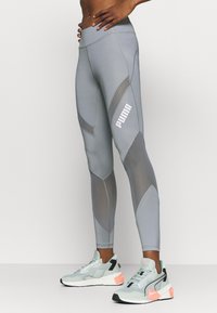 Leggings atletici grigi con pannelli a texture contrastante, dotati di vita alta e un prominente logo bianco Puma. Abbinati a scarpe da ginnastica verde menta.