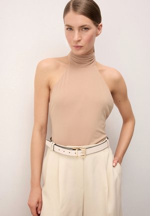 Femme aux cheveux lisses tirés en arrière portant un haut beige sans manches à col licou, rentré dans un pantalon crème taille haute avec une ceinture blanche.