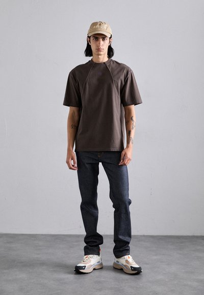 Brown cotton t-shirt with short sleeves, dark denim jeans, and beige cap. Footwear features a chunky sole with multi-colored accents.