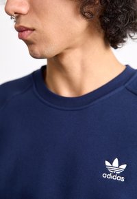 Mørkeblå sweatshirt lavet af bomuldsblanding, med ribbet rund halsudskæring og hvidt broderet adidas-logo på venstre bryst.