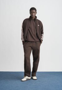 Chándal atlético marrón con detalles en rayas rosas, que incluye una chaqueta con cremallera y pantalones a juego, combinado con zapatillas blancas y negras.