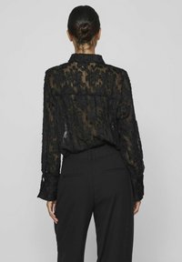 Blouse noire transparente avec texture florale, manches longues et col à revers. Associée à un pantalon noir cintré. Vue de dos.