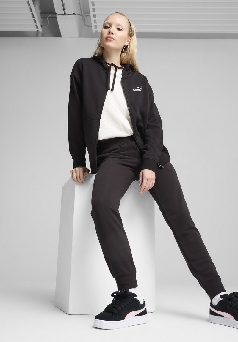Haut de sweat à capuche noir zippé avec logo, t-shirt blanc, pantalon de survêtement noir et baskets noires avec des accents roses, assis sur un bloc blanc.