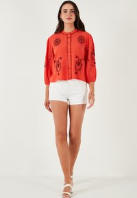 Rote Bluse mit schwarzer Stickerei, runder Ausschnitt, Knöpfe entlang der Vorderseite und Puffärmel. Kombiniert mit weißen Shorts und Riemchensandalen.