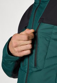 Padded jakna ima tirkizno tijelo s crnim detaljima, džepove na patentni zatvarač i glatku teksturu. Ruka prilagođava patentni zatvarač.