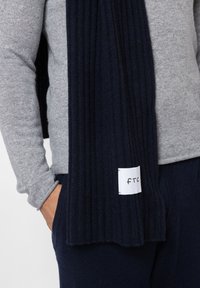 Marine ribbel sjaal gemaakt van zacht materiaal, met een wit label met "ftc." Dragen met een grijze trui, die de textuur en kleurcontrast toont.