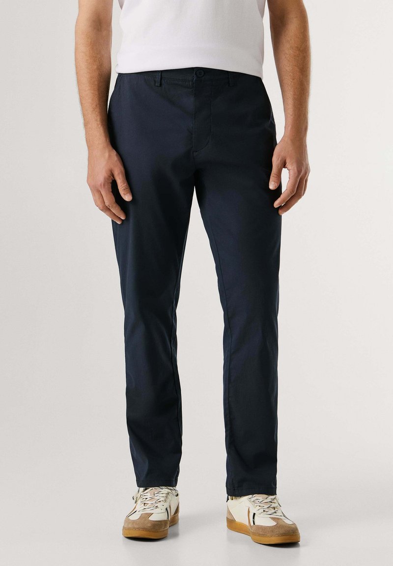 Homme portant un pantalon slim bleu marine foncé, des baskets blanches avec des détails beige et noir, et une chemise blanche rentrée, debout sur un fond uni.