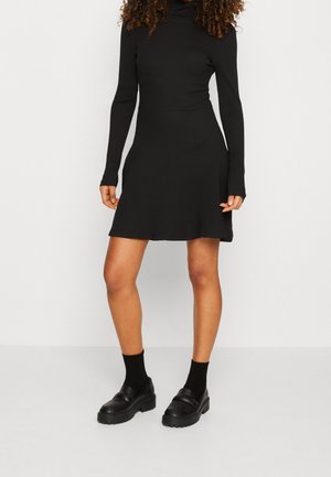 Femme portant une robe noire côtelée à manches longues et col roulé, associée à des chaussettes noires et des mocassins noirs épais, debout devant un fond blanc.