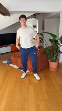 Sötétkék joggingnadrág világoskék logóval, világosszürke pólóval és fehér tornacipővel párosítva, fapadlón, egy cserepes növény közelében.