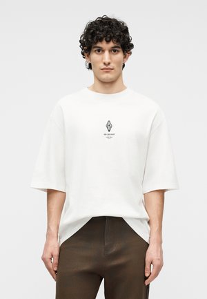 Mladý muž s kučeravými tmavými vlasmi, oblečený v bielej veľkej tričku s malým geometrickým logom a tmavými nohavicami na jednoduchom pozadí.