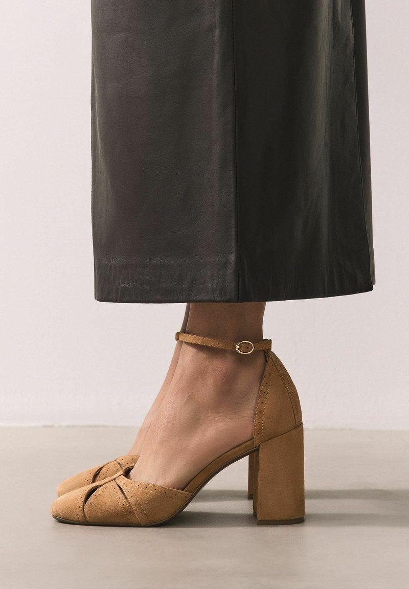 Beige Wildleder-Absatzschuhe mit Blockabsatz und Knöchelriemen. Verfügen über ein Cutout-Design und perforierte Akzente für zusätzliche Textur.