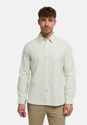 Mężczyzna w jasnorozowej i białej paskowanej koszuli na guziki oraz beżowych spodniach, stojący na jednolitym jasnym tle.