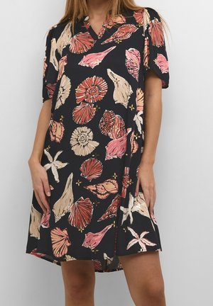 Frau trägt ein schwarzes Kleid mit kurzen Ärmeln und Muschel- sowie Seesternmustern in Rot-, Beige- und Weißtönen.