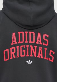 Čierna mikina s kapucňou, na ktorej je výrazný červený text "ADIDAS ORIGINALS" a biele logo Adidas trefoil. Materiál má hladký, matný povrch.