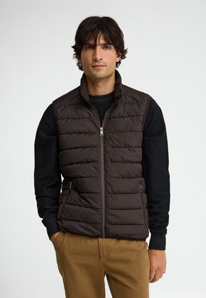 Uomo che indossa un gilet nero trapuntato senza maniche sopra una camicia nera a maniche lunghe e pantaloni marroni, in piedi davanti a uno sfondo bianco uniforme.