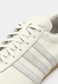 Λευκό δερμάτινο αθλητικό παπούτσι με τρεις ανοιχτό γκρι ρίγες από τσόχα, επίπεδα λευκά κορδόνια και υφασμένο καφέ πέλμα. Διαθέτει ραφές με λεπτομέρειες.