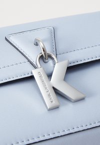 Světle modrá kožená kabelka s trojúhelníkovým klopou, stříbrným kováním a kovovým přívěskem ve tvaru písmene 'K'. Má viditelné švy podél okrajů.