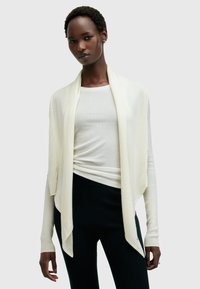 Leichte, elfenbeinfarbene Strickjacke mit offenem Frontbereich, gerippten Bündchen und weicher Textur, getragen über einem enganliegende cremefarbenen Oberteil und schwarzen Hosen.