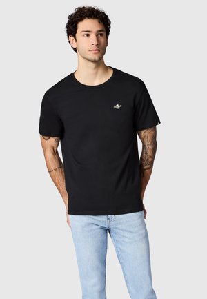 Man met zwart T-shirt met klein planeetlogo op de borst en lichtblauwe spijkerbroek, staand tegen een effen achtergrond.