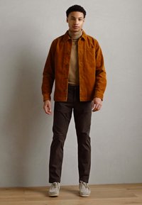 Brown corduroy shirt jacket over a beige turtleneck, paired with dark brown pants and light sneakers, against a neutral backdrop.