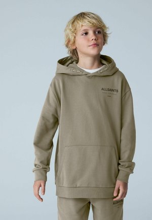 Lyshåret dreng iført en beige AllSaints-hoodie og matchende bukser, stående med et neutralt udtryk og kigger let mod højre.